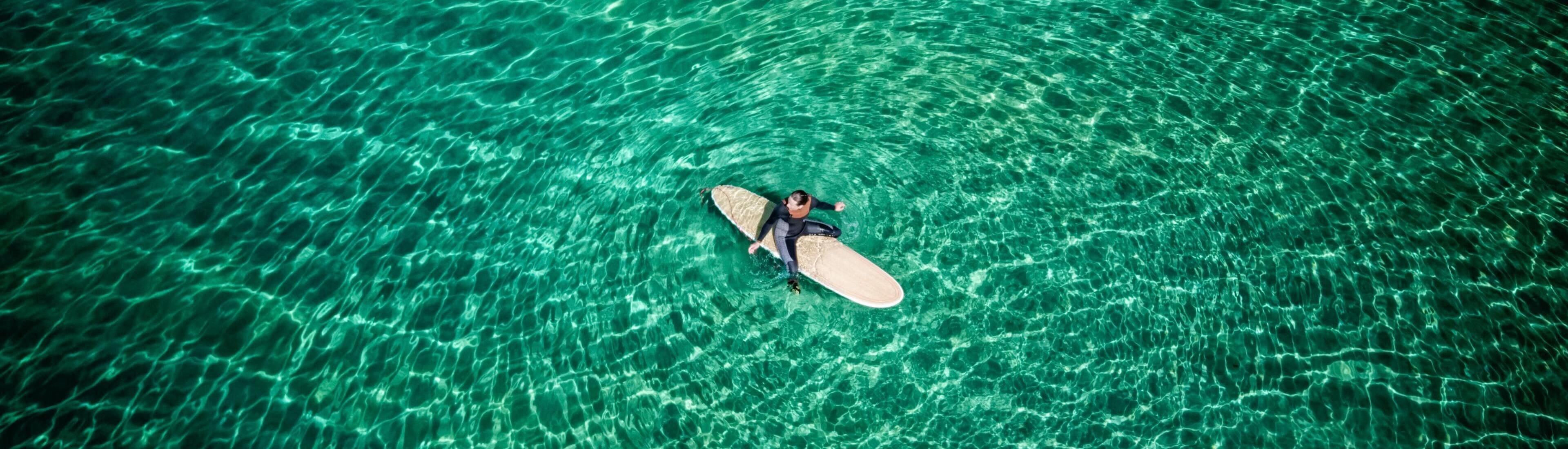 Person sitting on a long surfboard in green, calm water, viewed from above
